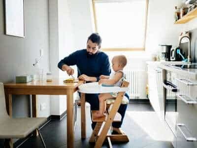 Father feeding a toddler in a high chair at a modern kitchen table, bright sunlight coming through a window.