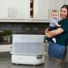 Tommee Tippee Super-Steam Advanced Electric Steriliser in a kitchen setting with a woman holding a baby in the background.