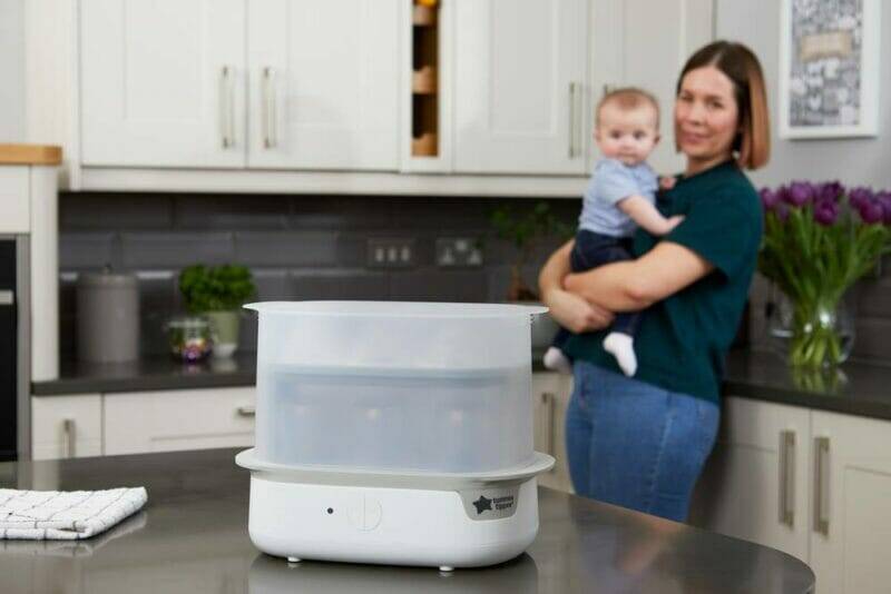 Tommee Tippee Super-Steam Advanced Electric Steriliser in a kitchen setting with a woman holding a baby in the background.
