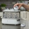 Tommee Tippee Super-Steam Advanced Electric Steriliser in white, sterilising multiple baby bottles with a hand placing a lid.