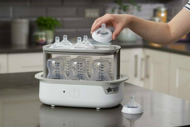 Tommee Tippee Super-Steam Advanced Electric Steriliser in white, sterilising multiple baby bottles with a hand placing a lid.