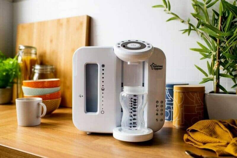 Tommee Tippee Perfect Prep Machine in white, placed on a wooden kitchen counter with colorful bowls and plants in the