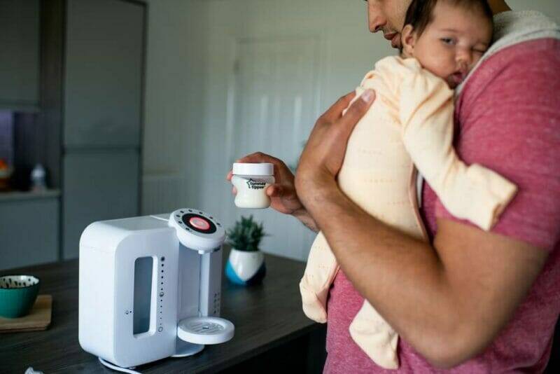 Tommee Tippee Perfect Prep Machine in white, with a parent holding a baby and a bottle, set in a modern kitchen.