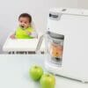 Tommee Tippee Quick-Cook Baby Food Maker in white, with fresh fruit in the bowl and a baby in a high chair.