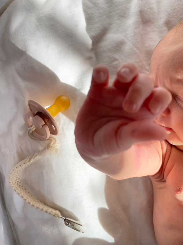 Baby's hand reaching out next to a beige pacifier with a yellow nipple and a cream-colored pacifier clip on a white blanket.