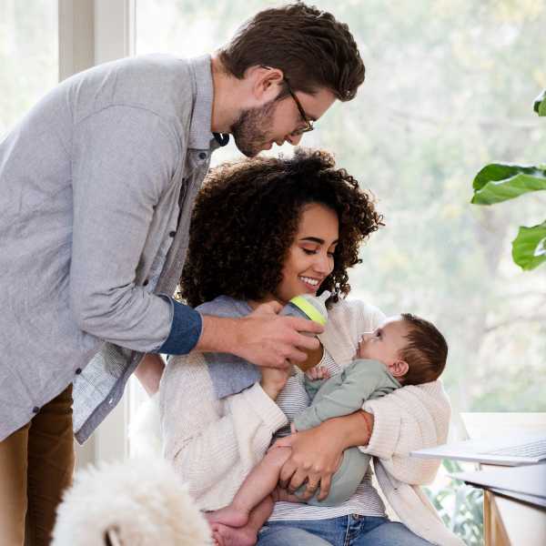 Couple feeding their baby with a bottle while sitting in a bright, cozy room with plants and a dog nearby.