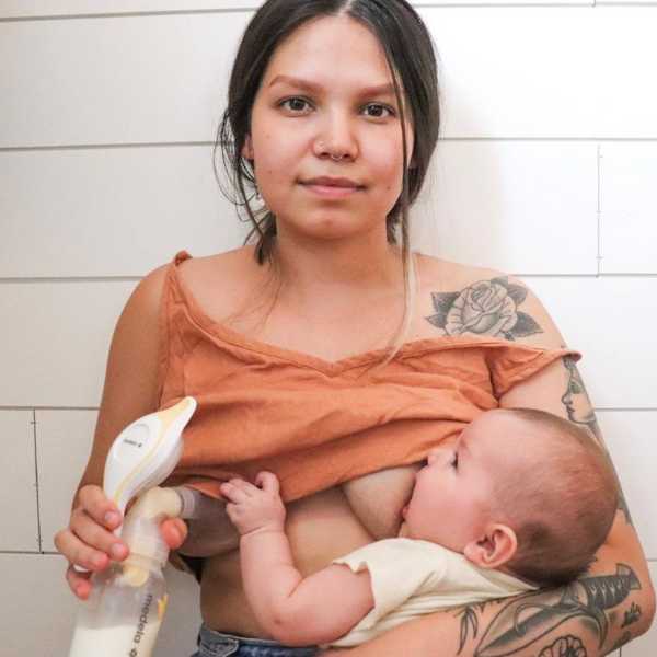 Mother breastfeeding her baby while holding a breast pump, wearing an orange top, with a wooden wall background.