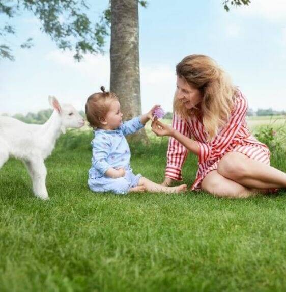 Mother and child sitting on grass, smiling and interacting, with a playful goat nearby in a sunny outdoor setting.