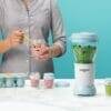 Nutribullet Baby blender in light blue with green peas inside, person using a spoon to serve puree from a glass jar.