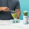 Nutribullet Baby in light blue, blending green puree with a smiley cup, set on a marble countertop against a turquoise