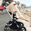 The Stella Clip in Beach Bébé, a beige clip on a stroller holding a cozy blanket, set against a coastal promenade.