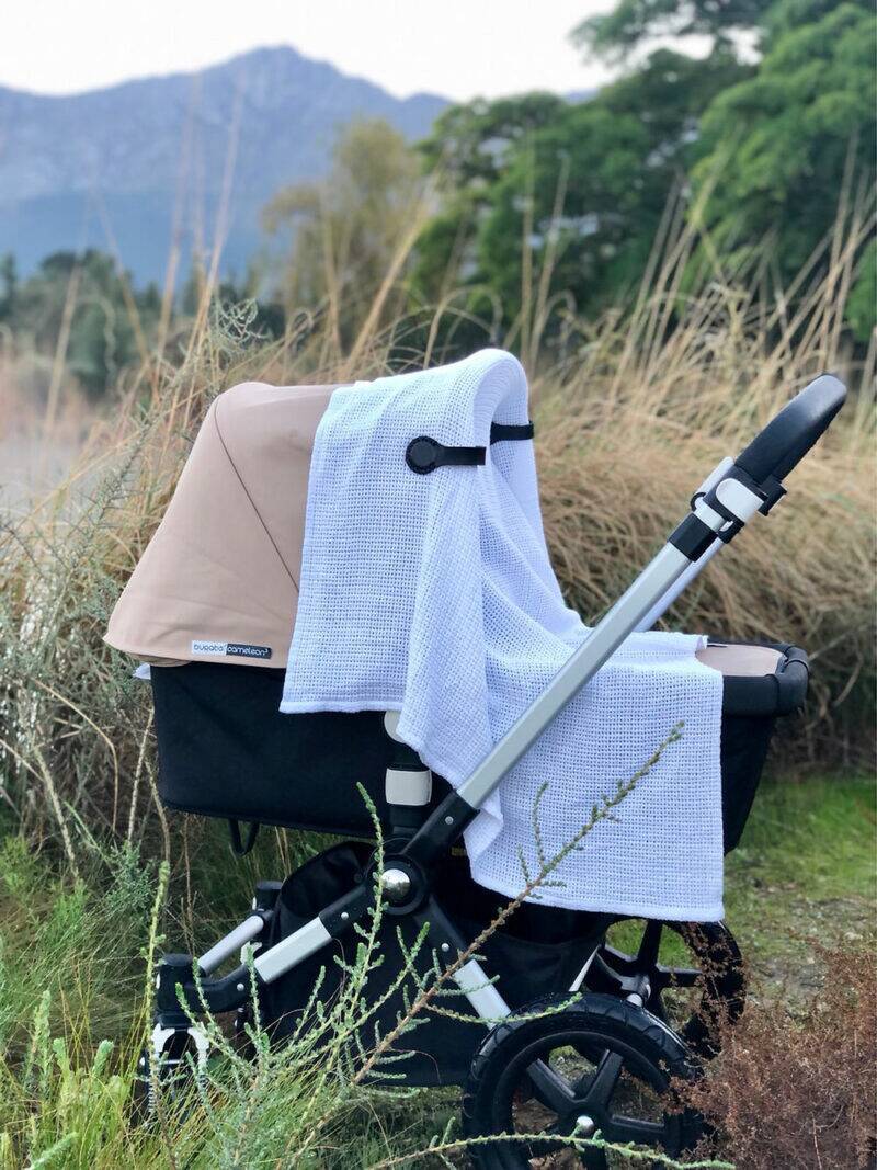The Stella Clip in Beach Bébé, a beige accessory draped over a stroller, set against a natural landscape with mountains.