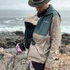 A man wearing a beige and teal jacket stands on rocky shore, holding a coffee cup and a baby in The Stella Clip carrier.