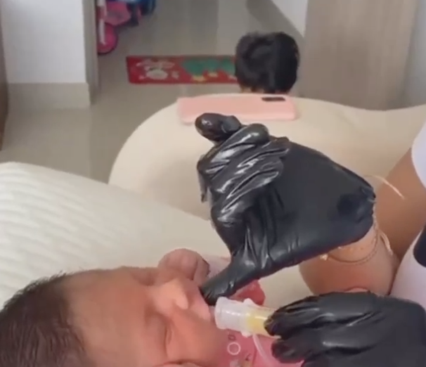 Baby being fed with a bottle by a caregiver wearing black gloves, in a cozy indoor setting.