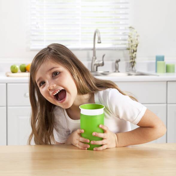 Munchkin Miracle 360° Sippy Cup in green, held by a smiling girl in a bright kitchen setting.