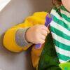 Munchkin Colour Reveal Toddler Forks & Spoons Set in purple, held by a child wearing a yellow sweater and striped bib.