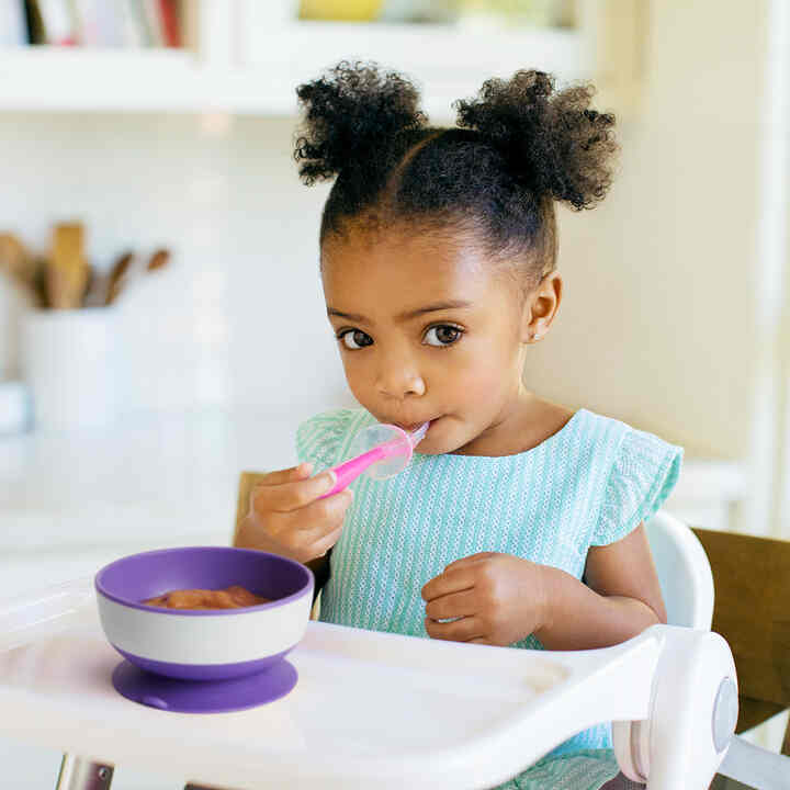 Munchkin Gentle Scoop™ Silicone Training Spoon in pink, used by a toddler in a high chair, enjoying food from a bowl.