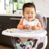 Munchkin C'Est Training Cup with straw in a toddler's hand, sitting in a high chair, smiling while enjoying a drink.