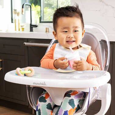 Munchkin C'Est Training Cup with straw in a toddler's hand, sitting in a high chair, smiling while enjoying a drink.