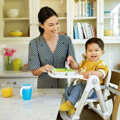 Munchkin Raise Toddler Fork 'n Spoon Set in blue, green, pink, and purple, with a happy toddler using them in a high chair.