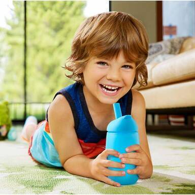 Munchkin Simple Clean Straw Cup in blue, held by a smiling boy lying on a colorful rug in a bright room.