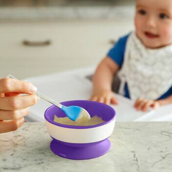 Munchkin Stay Put Suction Bowl in purple and white, held with a spoon, with a baby in a high chair in the background.