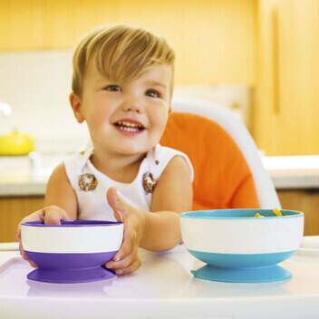 Munchkin Stay Put Suction Bowls in purple/green/blue and yellow/green/blue, held by a smiling toddler in a high chair.