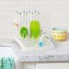 Munchkin Deluxe Bottle Drying Rack in white with green and clear accessories, set on a marble countertop with colorful bowls.