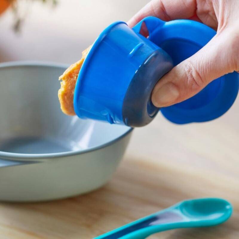 Tommee Tippee Pop Up Freezer Pots in blue being used to release food into a gray bowl, with a teal spoon nearby.