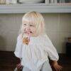 Child playing with FRIGG + Mushie Nature Teething Ring in a cozy kitchen setting, wearing a white blouse and smiling.