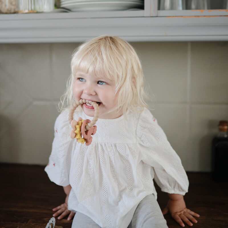 Child playing with FRIGG + Mushie Nature Teething Ring in a cozy kitchen setting, wearing a white blouse and smiling.
