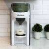 Baby Brezza Formula Pro Advanced in black, dispensing formula into a clear cup on a kitchen counter with potted plants.