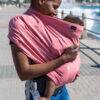 Maccie Stretch Cover in Ruby Pink, worn by a woman holding a baby, with a scenic waterfront in the background.