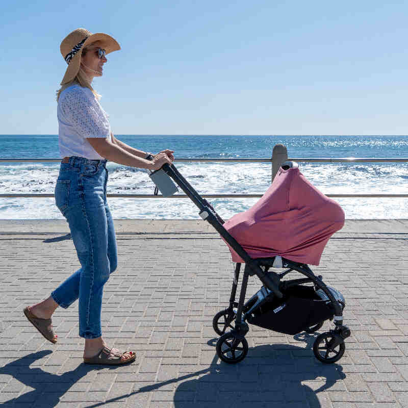 Maccie Stretch Cover in Ruby Pink draped over a stroller, with a woman walking by the beach in casual attire and a sun hat.