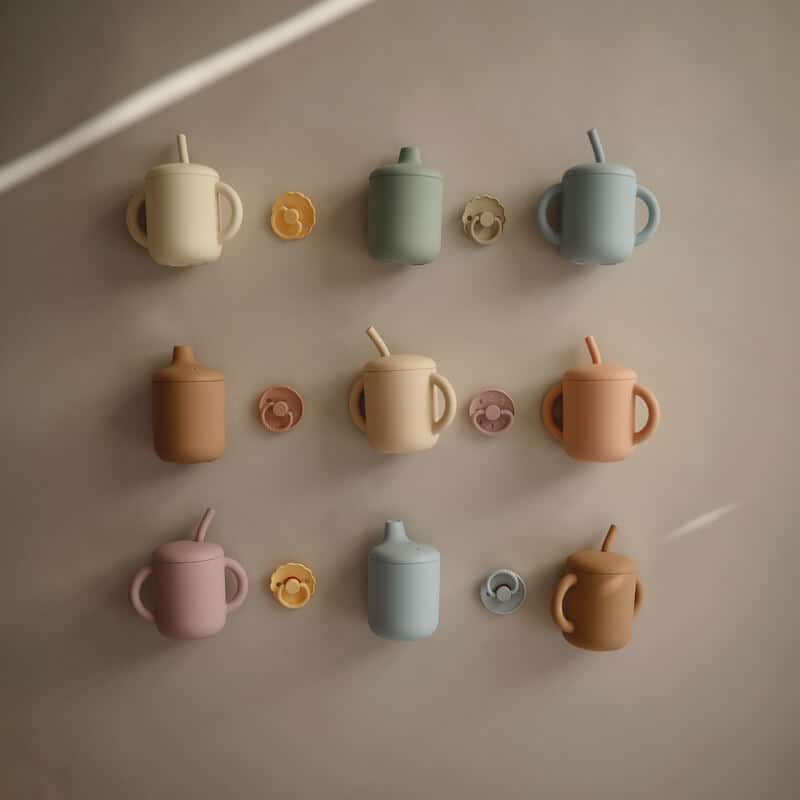 Mushie Silicone Training Cups in various colors displayed on a wall, featuring straws and matching lids.