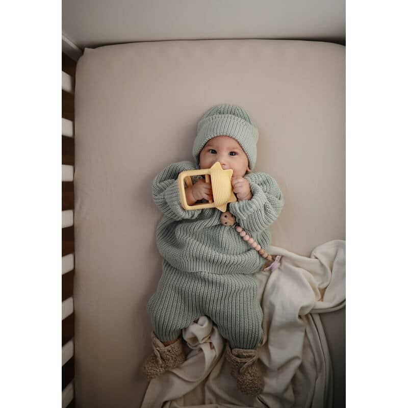 Mushie No-Drop Sun Teether in Sunshine, held by a baby in a cozy outfit, lying on a soft blanket in a crib.