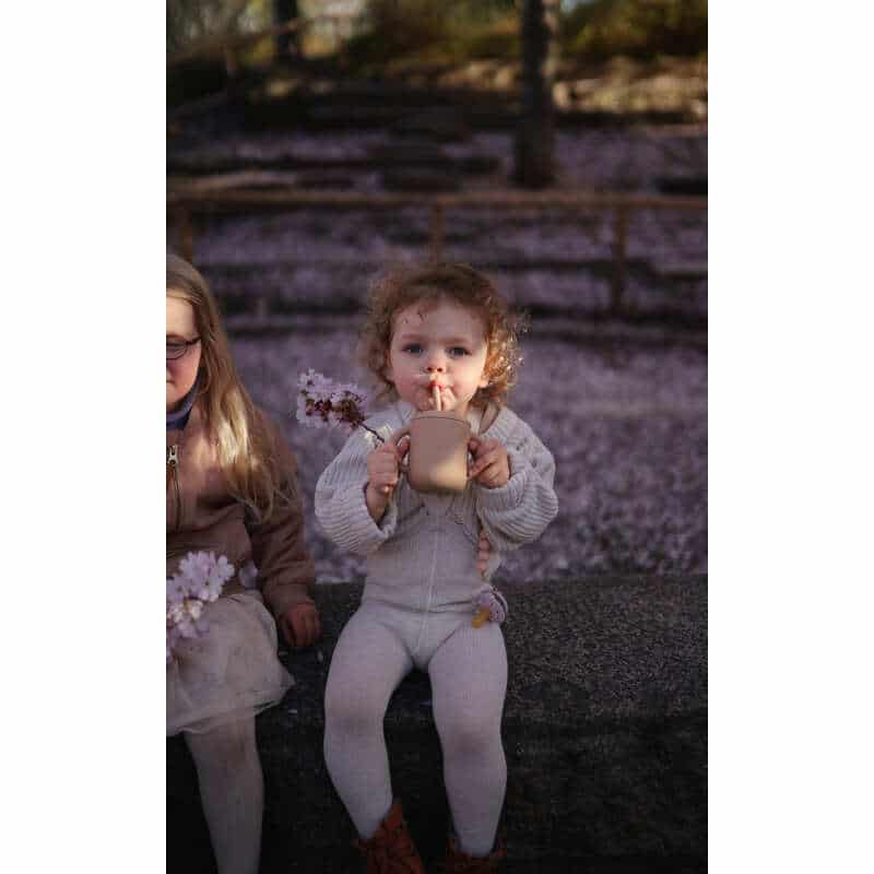Mushie Silicone Training Cup in Blush, held by a child outdoors, surrounded by cherry blossoms, showcasing its playful