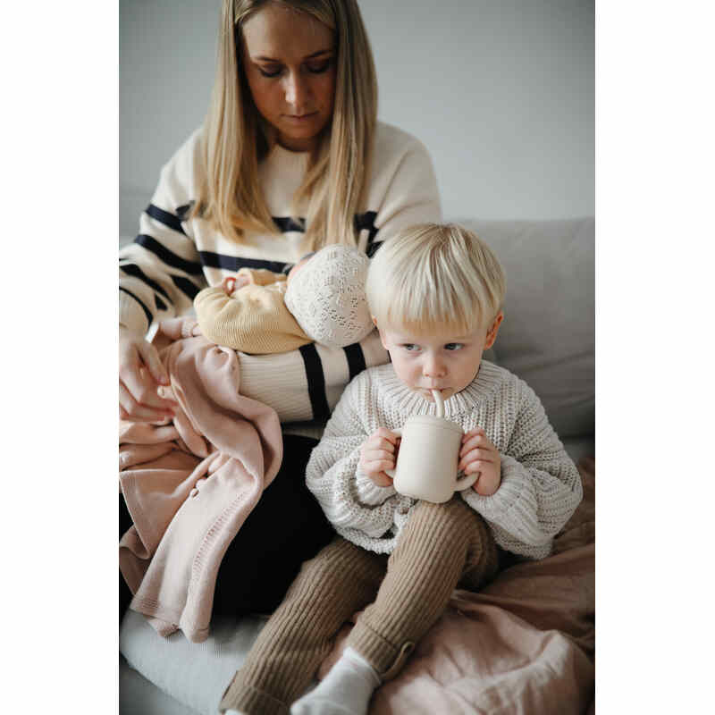 Mushie Silicone Training Cup in Blush held by a child sitting next to a parent, who is holding a baby. Cozy indoor setting.