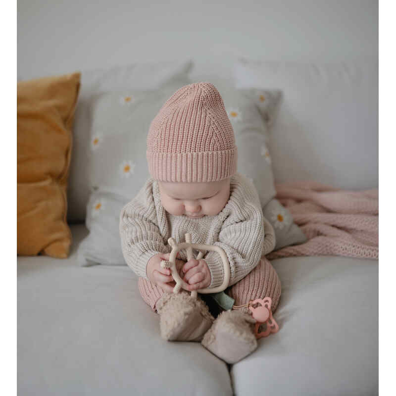 Mushie No-Drop Sensory Teether in Blush, held by a baby sitting on a couch surrounded by soft pillows and a blanket.