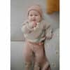 Mushie No-Drop Sensory Teether in Blush, held by a baby in cozy knitwear, lying on a soft couch with decorative pillows.