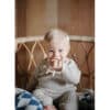 Mushie No-Drop Sensory Teether in blush and shifting sand, held by a smiling baby in a cozy, woven chair setting.