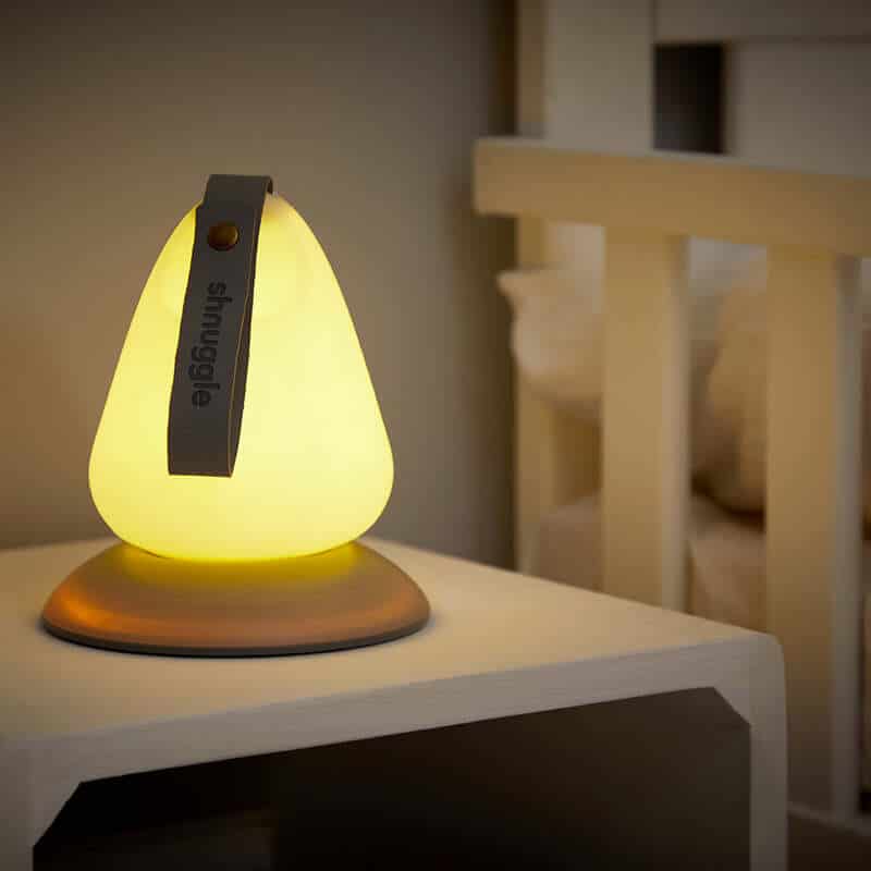 Shnuggle Moonlight Night Light in soft yellow, shaped like a teardrop, placed on a bedside table next to a crib.
