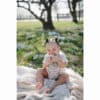 Mushie Silicone Fresh Food Feeder in blush, used by a smiling toddler outdoors on a blanket surrounded by flowers.