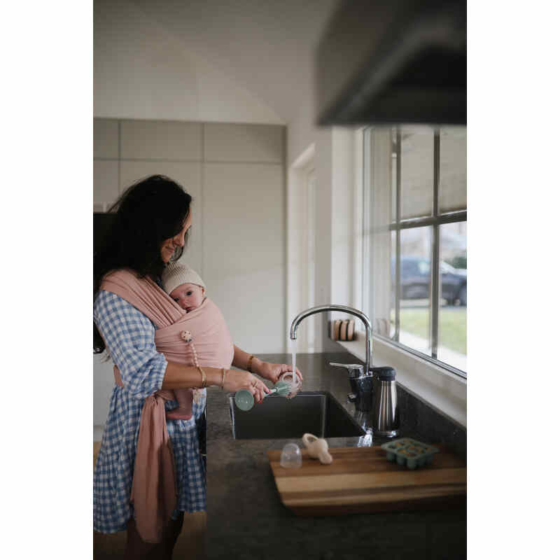 Mushie Silicone Baby Bottle Brush in blush, held by a mother in a kitchen while caring for her baby.