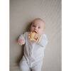 Mushie No-Drop Sun Teether in Sunshine color held by a baby wearing a white onesie, lying on a neutral background.
