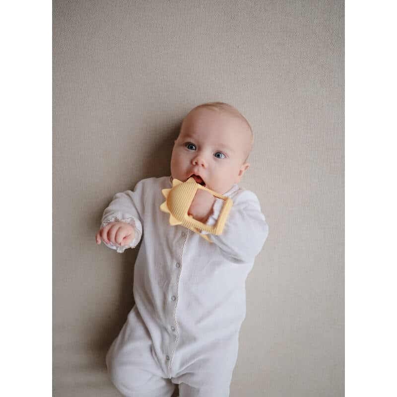 Mushie No-Drop Sun Teether in Sunshine color held by a baby wearing a white onesie, lying on a neutral background.