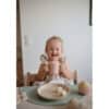 Mushie Silicone Training Cup in Blush, held by a smiling child at a dining table with a plate of fruit.