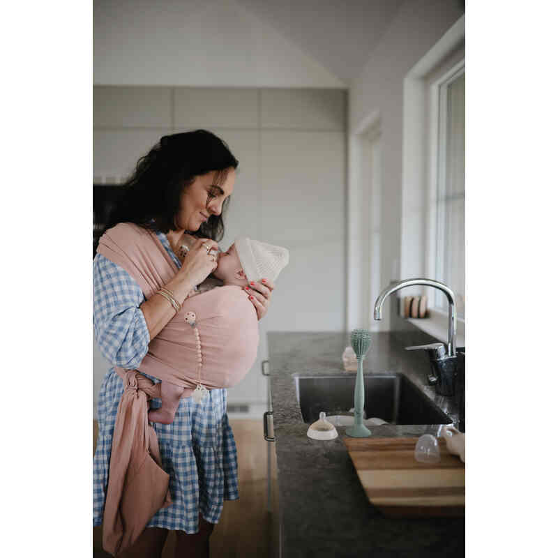 Mushie Silicone Baby Bottle Brush in blush, shown in a kitchen setting with a mother holding her baby in a wrap.