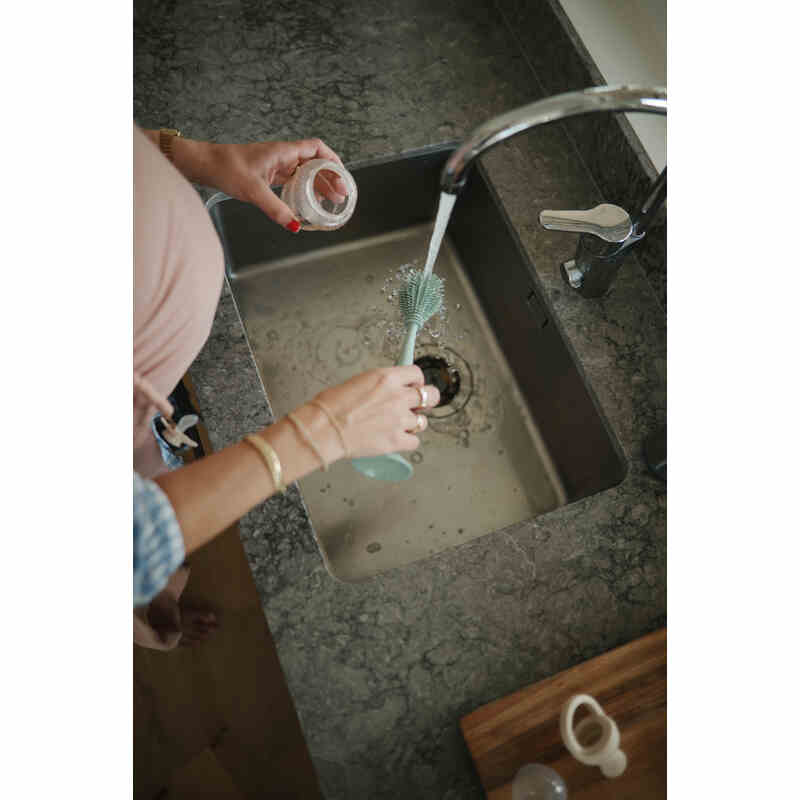 Mushie Silicone Baby Bottle Brush in Cambridge Blue being used in a sink, with hands holding a bottle and brush.