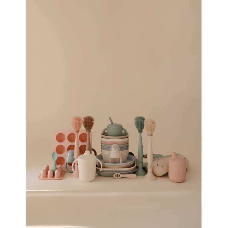 Mushie Silicone Baby Bottle Brush in blush among various baby utensils and dishes in soft pastel colors on a neutral
